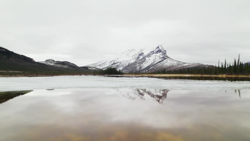 An icy lake mirrors the snowy mountains in the distance