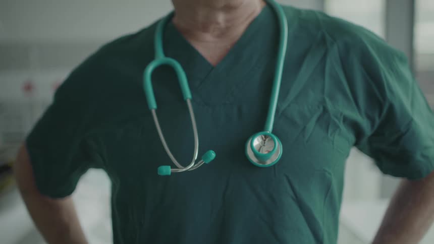 A doctor with scrubs and a stethoscope in a room with a wall in the background