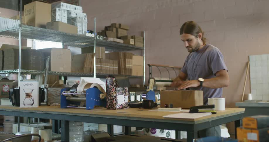 A man in a warehouse tapes a cardboard box closed and carries it out of frame