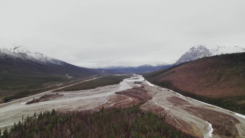 Snow and ice cover partly dry riverbed bordered by mountains hills and forest under clouded sky