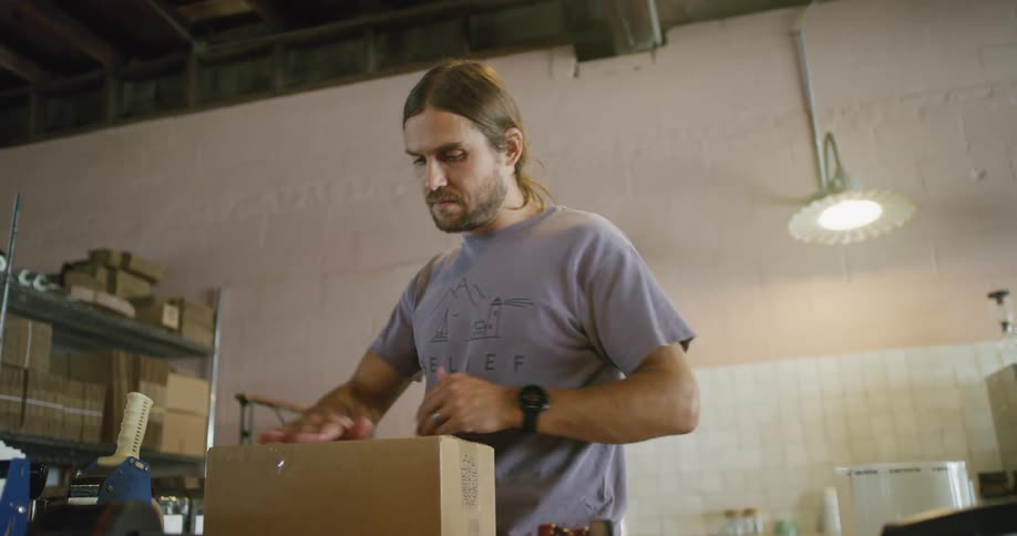 A man tapes a box closed before carrying it out of frame