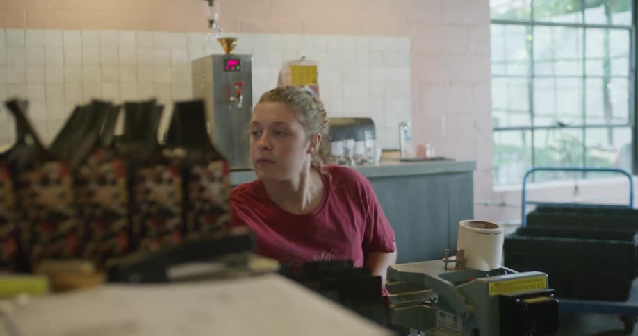 Fair haired woman binds packages of coffee with coffee roaster and grinder in the background
