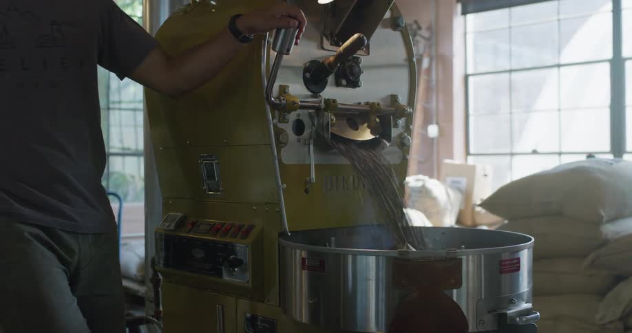 A man in a room pulling a lever on a machine that dispense out coffee beans with a wall and a window in the background