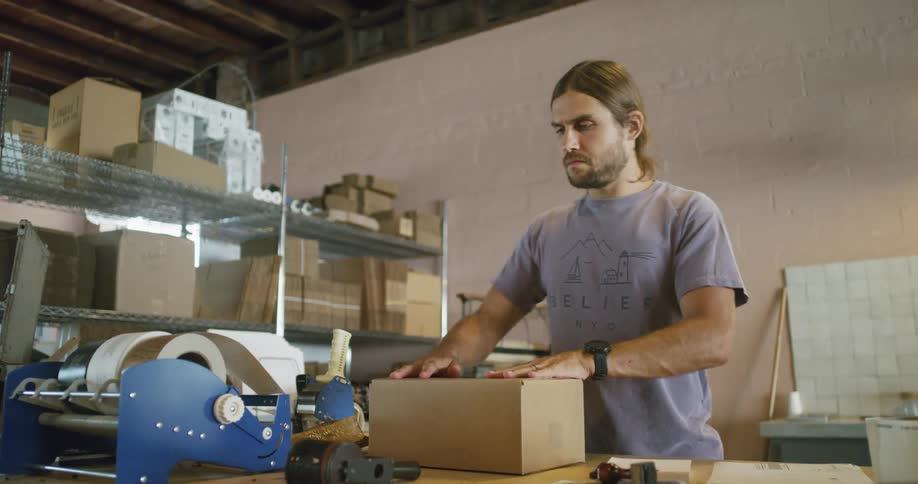 A man places paper into a cardboard box then pulls tape to seal the box for shipping