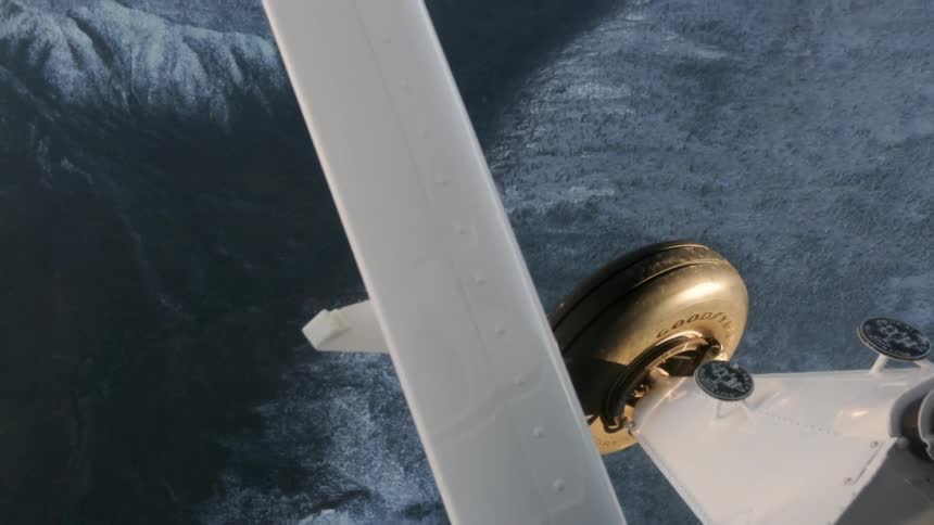 The landing gear of a small plane are seen as it flies over a snow-covered mountain range