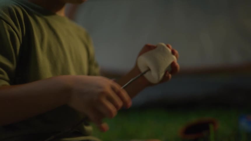 A man sits outside beside a boy who puts a marshmallow on a stick