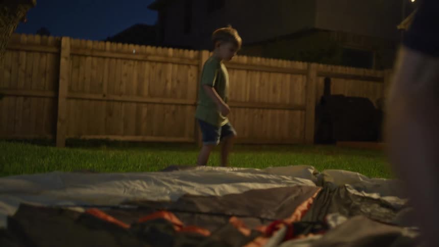 A man and his small son assemble a tent in the backyard of their house at night