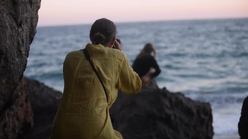 A woman takes a photograph of a woman sitting on a rock watching the ocean waves at sunset