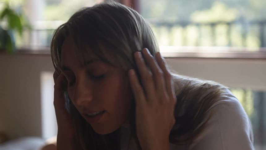 A woman sitting on a porch with her head bowed pushes her hair back from her face