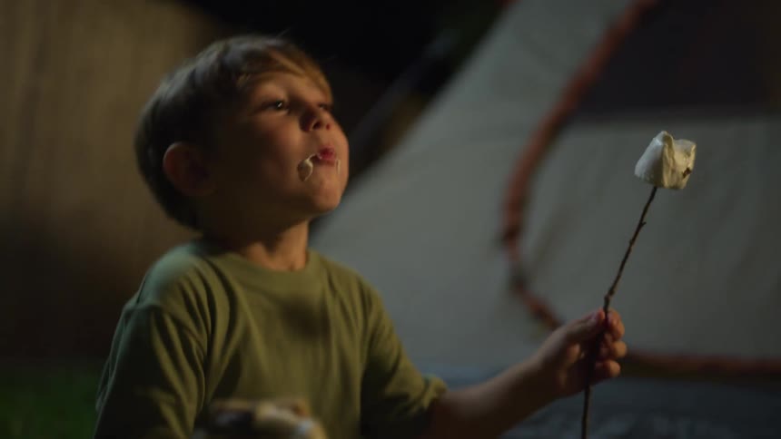 A young boy eating a roasted marshmallow from a stick while camping