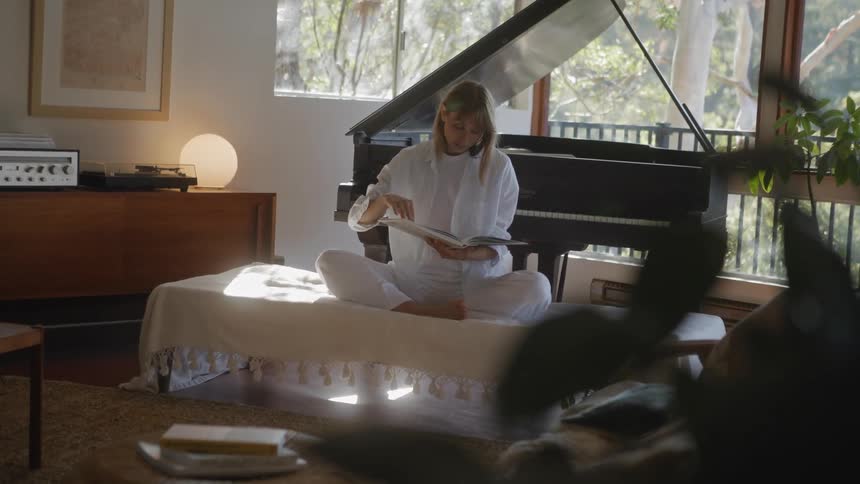 A woman sits and reads on a bench in her living room in front of a piano