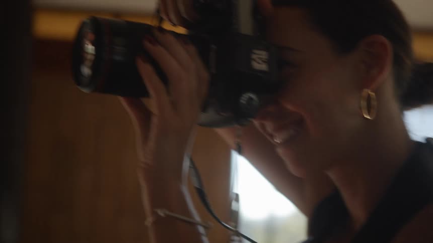 A photographer holds a professional camera up to her face taking pictures inside a building with large windows