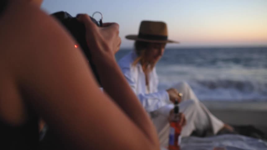 A woman sitting on a towel on the beach attempts to open a bottle of wine as a photographer takes a photograph of her