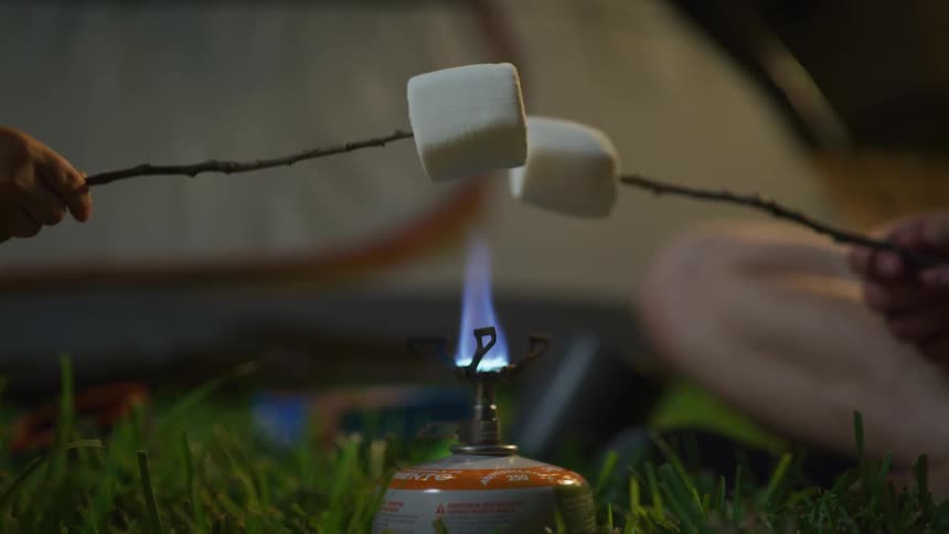 A pair of campers toast marshmallows over a burner while sitting in the grass outside their tent at night