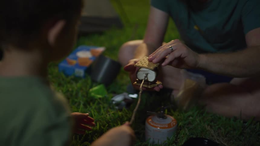 A man helps a boy to make a chocolate smore while camping at night