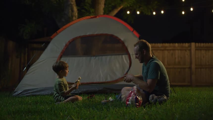 A dad and son sit in the backyard eating smores and marshmallows near their tent at night