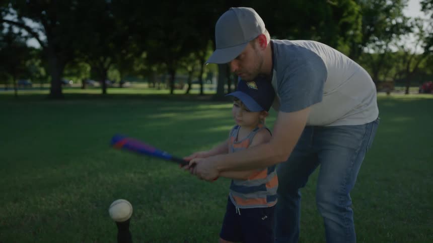 A man showing a boy how to hit a baseball with a baseball bat on a t with trees the sky and clouds in the background