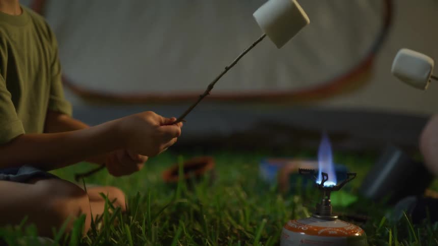 Kid sits outdoors on grass roasting large marshmallows next to tent