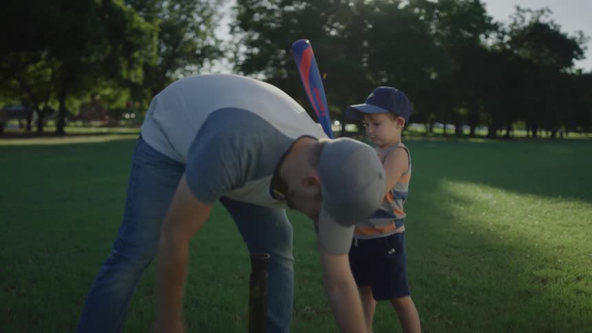 A man teaching a boy how to hit a baseball with a baseball bat on a t with trees the sky and clouds in the background