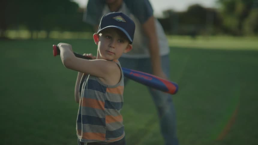 A young boy swings a baseball bat and starts running with his dad watching