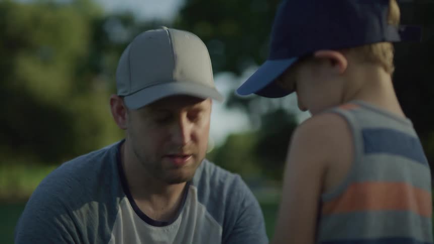 A man kneels down and talks to his young boy outside who then picks up a small baseball bat from the ground