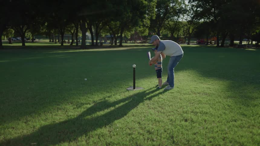 Father teaches his son how to hit a ball with a bat in a field on a sunny day