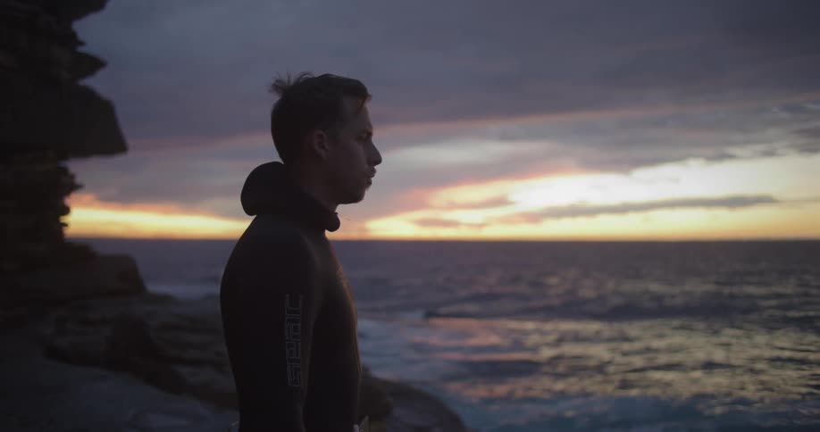 A man in a wetsuit standing on a rocky shore with the ocean sky and clouds in the background