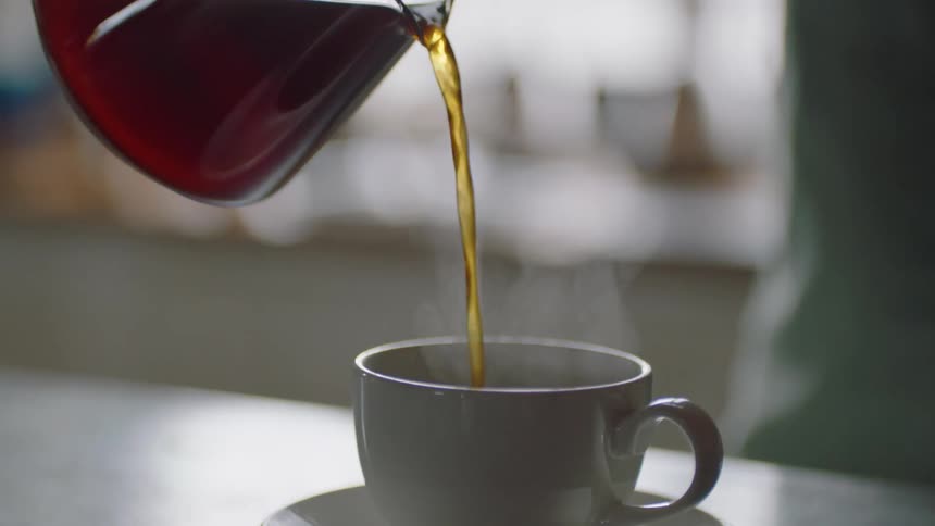 A person pouring hot coffee into a white mug on a saucer