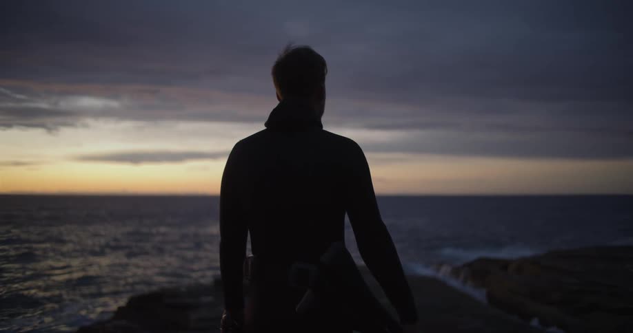 Silhouette of a man wearing a wet suit looking at the ocean at sunset