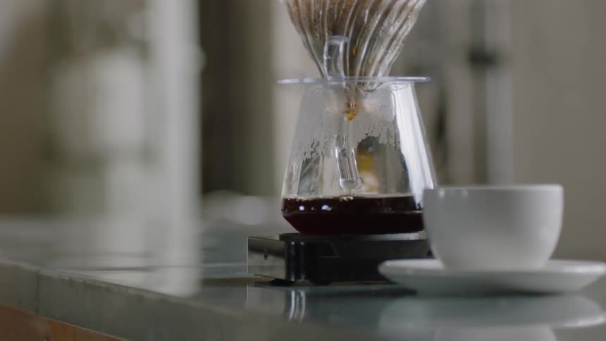 Coffee drips through filtered funnel into glass pitcher on counter next to white ceramic cup and saucer