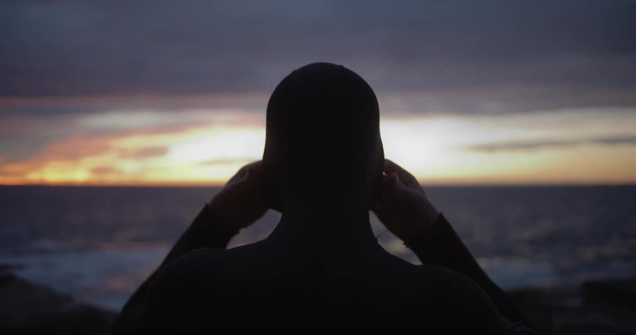 A man pulls the hood of his wet suit over his head while facing the horizon on the coast at sunset