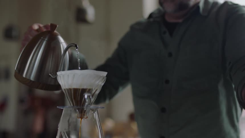 A man pours steaming water from a kettle into a coffee filter filled with coffee grinds causing coffee to drip into the coffee pot below