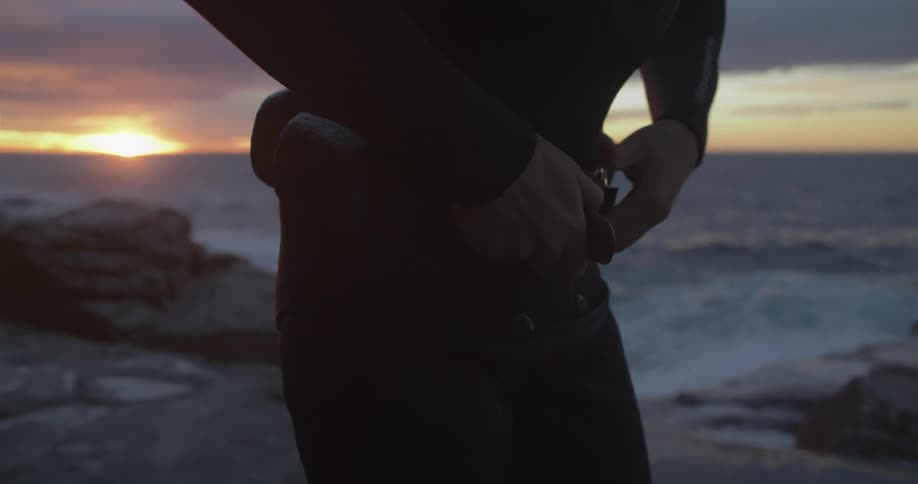 A man is putting on swimming gear by an ocean at dusk
