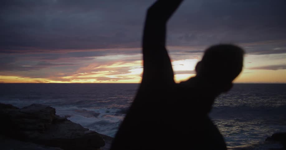 A silhouetted man stretching on a beach at sunset