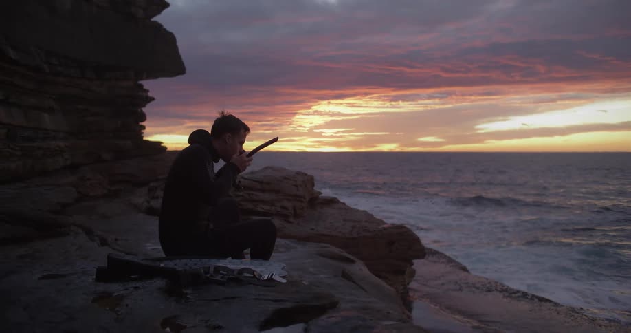 Man sits on a rock near the ocean and puts a snorkel up to his mouth