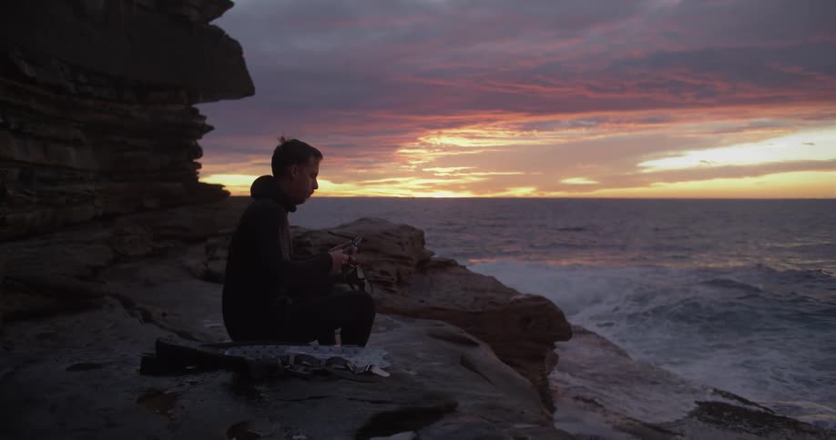 Young man wearing a black wet suit sitting on a promontory in front of the ocean at sunset