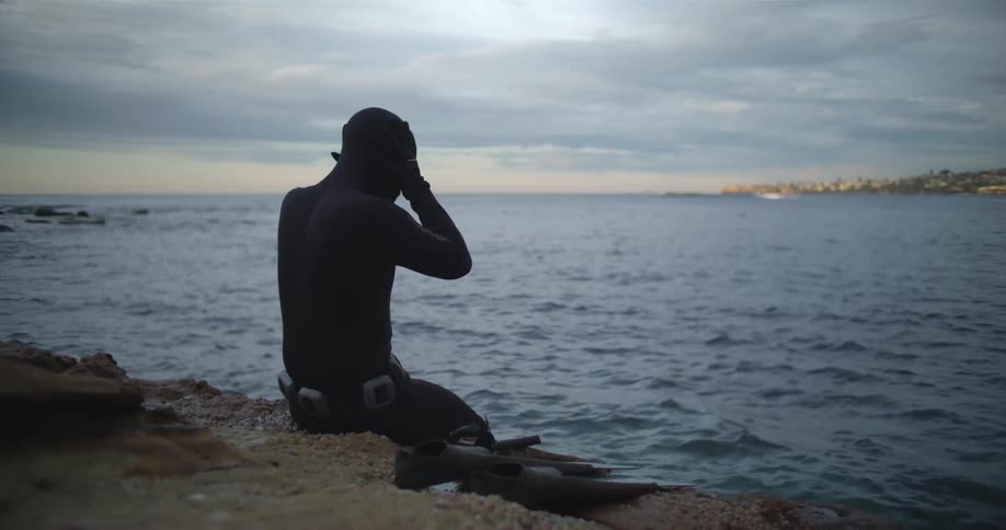 A man is sitting by the coast and putting on swimming gear