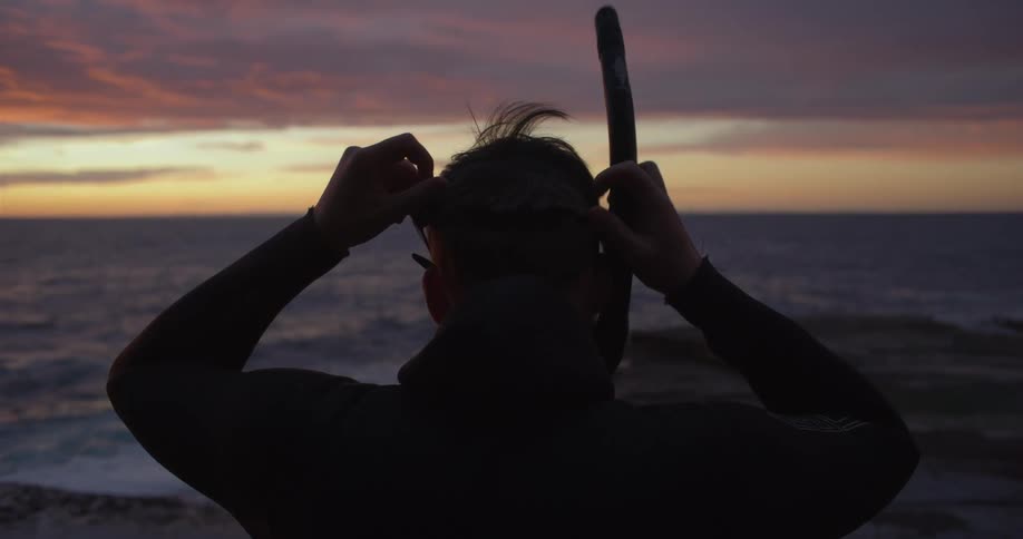 A diver puts on his diving mask while sitting on the rocky coastline at sunset