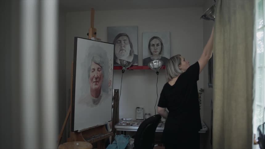 A woman turns on lights in a studio with many hanging paintings