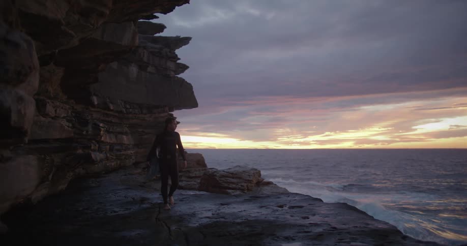 Man wearing a black wet suit carries a surfboard over a scarpment near the ocean at sunset