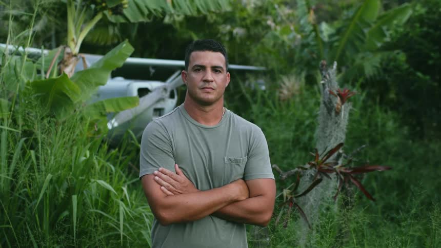 A man standing and staring with a airplane surrounded by plants and trees in the background