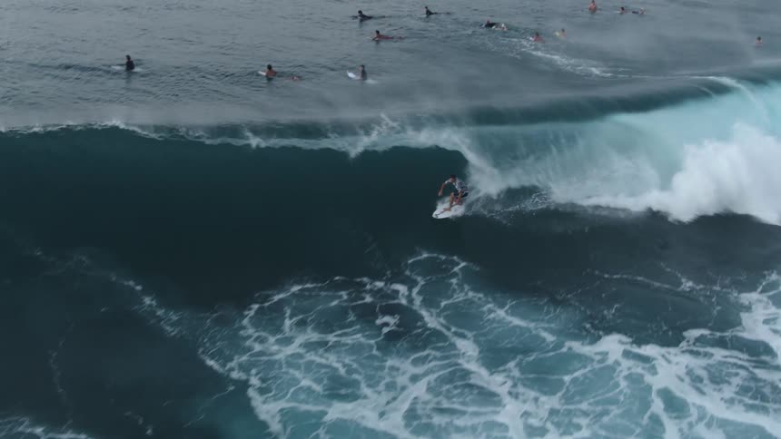Several surfers sit on their boards while one man rides a large wave
