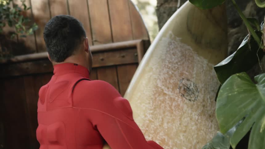 Man wearing a padded red wet suit opens a door in a wooden fence carrying a surfboard