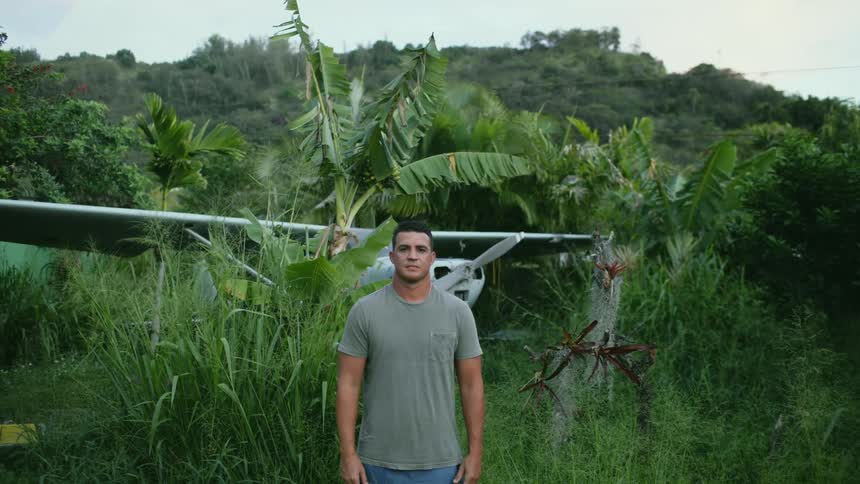 A man standing and staring with a airplane surrounded by plants and trees the sky and clouds in the background