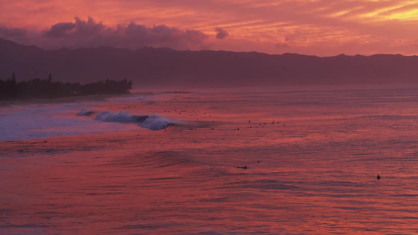 An aerial shot captures perfect waves breaking under a colorful red sky in Oahu, Hawaii