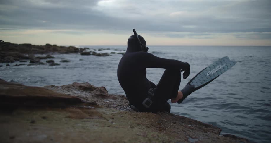 A person wearing a wet suit sits on a rocky beach and puts on flippers