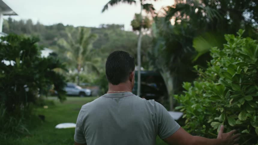 A man walking through a yard outdoors runs his hand through a bush as he passes