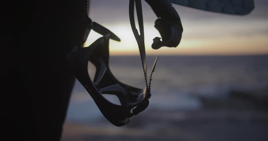 Person wearing wet suit and holding mask with snorkel stands on beach facing red tinted horizon over ocean