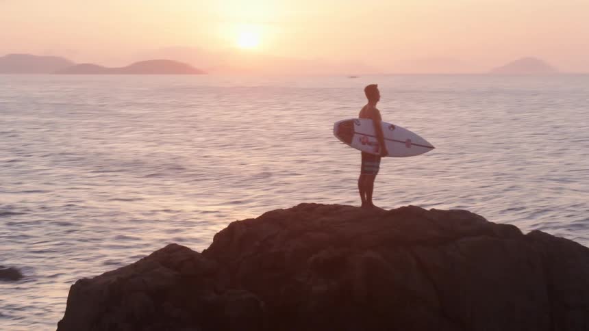 A man standing on a rocky coast holding a surfboard with the ocean sky and clouds in the background