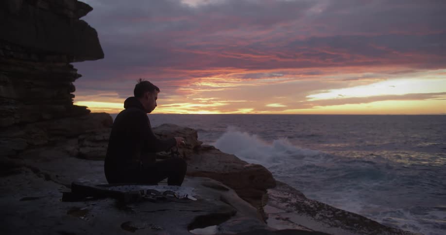 Silhouette of a man sitting on a beach watching the waves splash against the rocks as the sun is setting